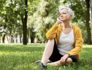 older woman in yellow sweater sits on grass. Meditating. Not crosslegged, just chin in hand.